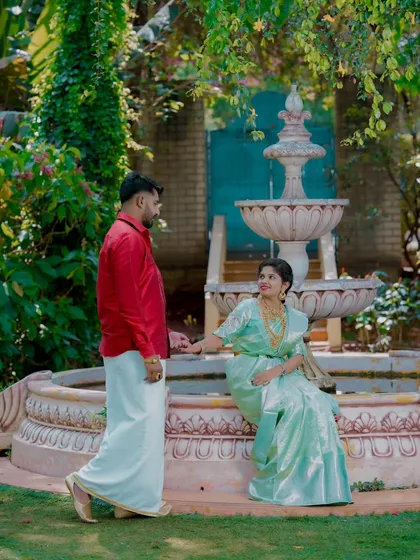 A romantic moment by a garden fountain. The bride's light green silk saree and the groom's red shirt and dhoti create a striking contrast.