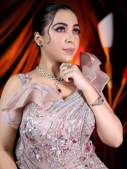 A glamorous studio shot of a model in a heavily embellished gown. The makeup is dramatic and high-fashion, with a sharp winged liner and pink eyeshadow.