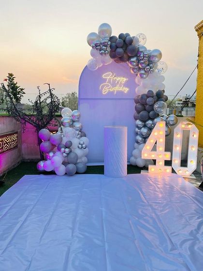 A 40th birthday celebration on a rooftop. This setup features a grey and white balloon arch against a custom backdrop, with a neon sign and marquee numbers.