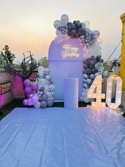 A 40th birthday celebration on a rooftop. This setup features a grey and white balloon arch against a custom backdrop, with a neon sign and marquee numbers.