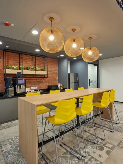 A pantry area with a high communal table and bright yellow chairs. The exposed brick wall and spherical pendant lights add warmth and texture.