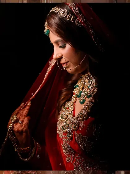 A dramatic and artistic bridal portrait. The use of shadow and light creates a timeless, painterly effect, emphasizing the bride's elegance and the richness of her red veil.