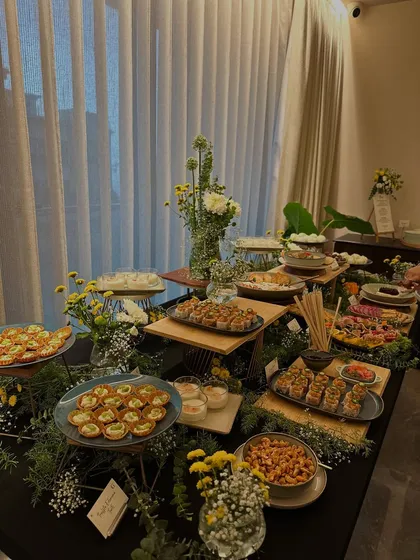 A wide shot of a beautifully styled grazing table against a curtained window. The multi-level display showcases a variety of appetizers, from tartlets to sushi, all nestled among fresh greenery.