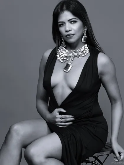 A seated black and white portrait in a plunging neckline dress, highlighting a bold statement necklace. The focus is on glamour and confidence.