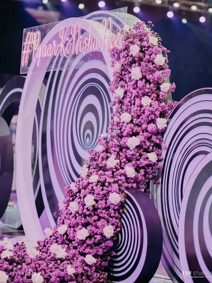 A neon sign with the couple's wedding hashtag, #PyaarKiNishaani, set against a cascade of purple flowers and a hypnotic spiral backdrop.