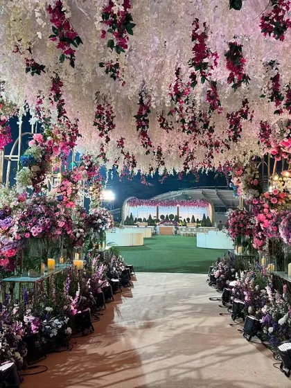 A magical walkway into an outdoor celebration. The path is lined with candles and lush floral arrangements, leading guests under a canopy of hanging flowers. 🩷💜