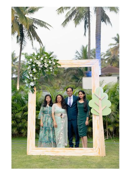 A family poses inside a large, rustic wooden photo frame we set up for an outdoor event. The frame is adorned with a corner floral piece, perfectly complementing the natural surroundings.