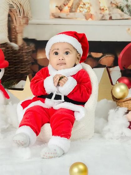 A baby dressed in a Santa suit sits like a little boss in a miniature armchair, surrounded by Christmas decorations.