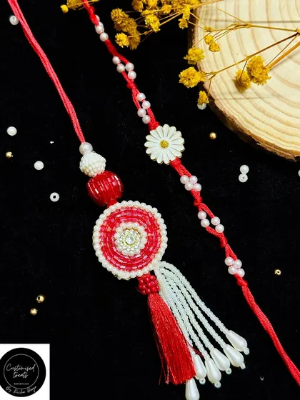 A vibrant red and white paired rakhi set. The Lumba features a circular beaded design with tassels, while the brother's rakhi has a delicate daisy charm.