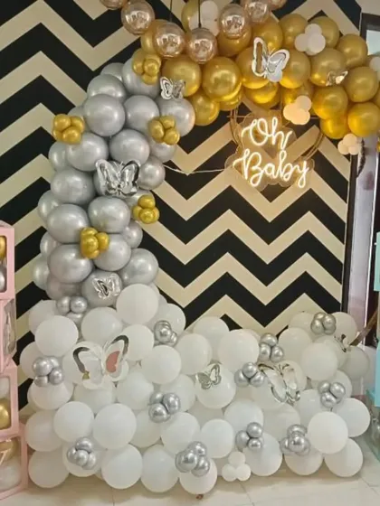 A chic baby shower setup with a half-moon balloon arrangement in white, silver, and gold against a bold chevron-patterned wall. The design is accented with butterfly cutouts and an "Oh Baby" neon sign.
