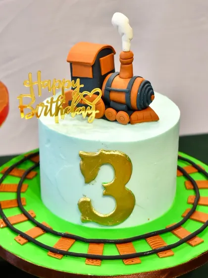 A close-up of one of the train-themed cakes, featuring a fondant steam engine on top and a large number 3, all set on a base of edible railroad tracks.