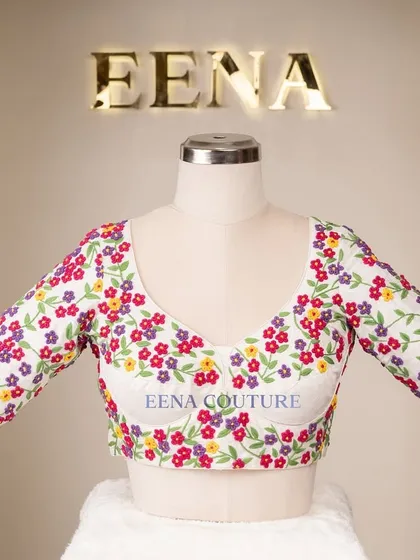 A garden of flowers on a blouse. This white raw silk piece is covered in vibrant, multi-colored floral French knot embroidery, making it a cheerful and eye-catching addition to any festive wardrobe.