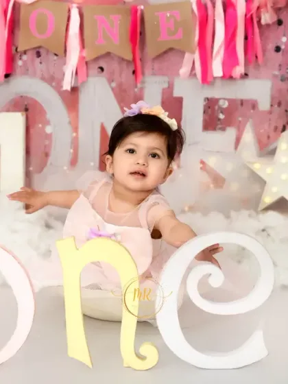 A baby girl sits among "One" letters and a pink, dreamy cloud setup for her first birthday celebration.