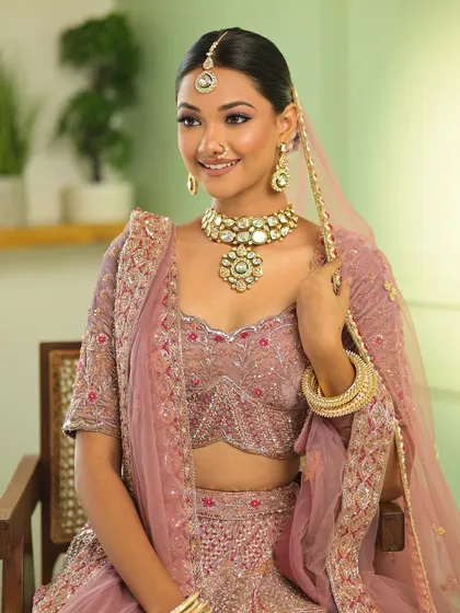 A full view of the bride seated, showcasing her beautiful pastel pink lehenga and the elegant, clean lines of her hair and makeup.