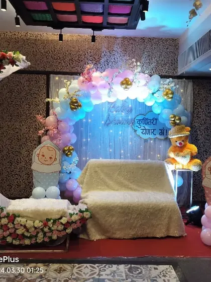 A whimsical baby shower setup featuring a large, floral-decorated moon cradle. The backdrop includes a balloon arch and a 'Kunitari Yenar Ga!' sign, creating a dreamy atmosphere for the mom-to-be.