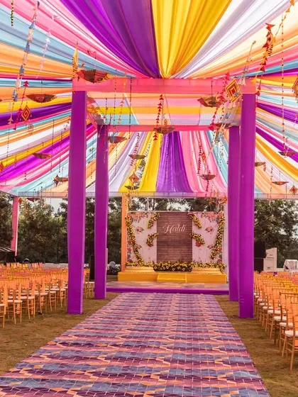 A full view of a vibrant Haldi ceremony setup, with a ceiling of multi-colored drapes and a spacious seating arrangement. I design these events to be comfortable and visually spectacular from every angle.