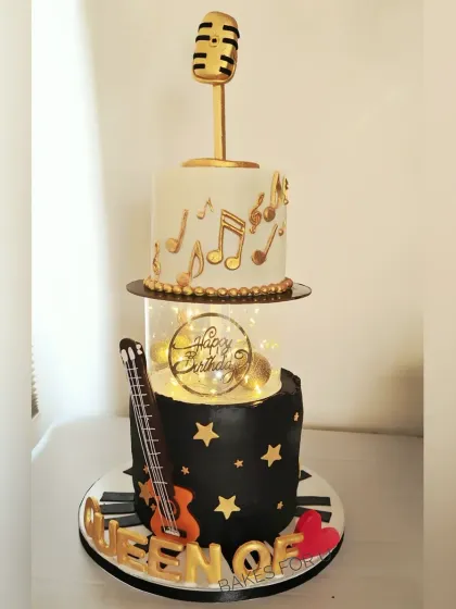 A two-tier music-themed cake for a "Queen of Fitness." The design combines a guitar, piano keys, a microphone, and fairy lights to celebrate her passions.