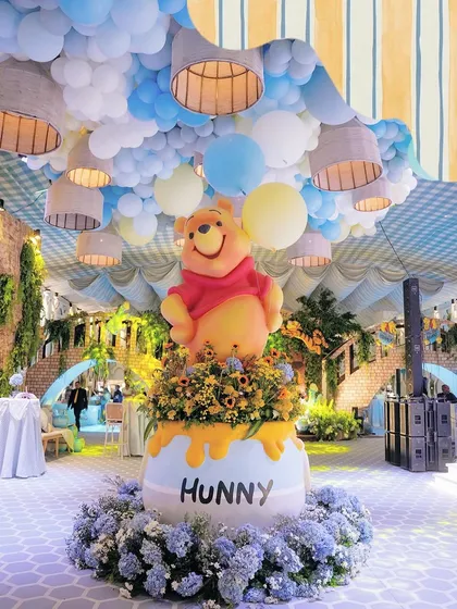 The centerpiece for a Winnie the Pooh first birthday, featuring a larger-than-life Pooh Bear atop a 'Hunny' pot overflowing with sunflowers. The ceiling is a sky of blue and yellow balloons, creating a storybook world.