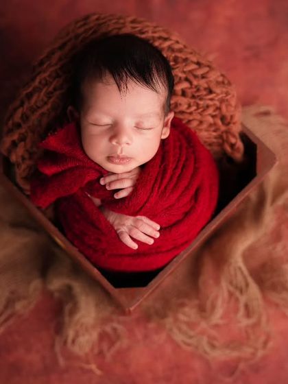 An overhead view of a newborn swaddled in a deep red wrap, resting peacefully inside a dark wooden box on a textured blanket.