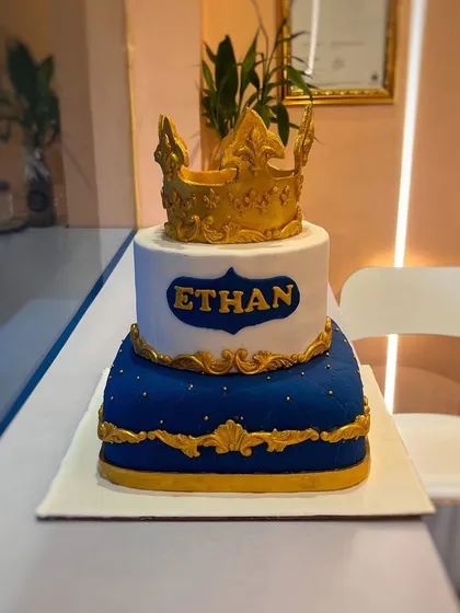 A cake fit for a prince. This two-tier creation features a royal blue quilted bottom tier and a white top tier, both with gold accents, and is topped with an ornate, handcrafted golden crown.