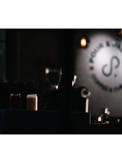 An atmospheric shot of the Pour & Palate cafe, with two drinks on the counter and the logo blurred in the background. The focus on the foreground creates a sense of depth and mystery.