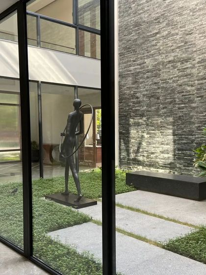 A minimalist courtyard in a New Delhi farmhouse, viewed through floor-to-ceiling glass. The space is defined by a striking modern sculpture, a bed of lush groundcover, and a simple stone bench, creating a living piece of art within the home.