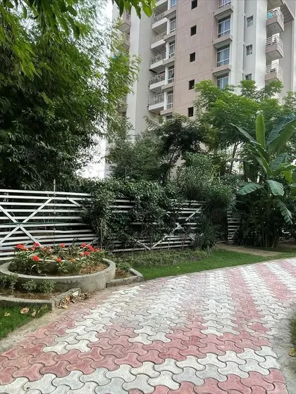 A corner of the park featuring a circular flower bed with red roses, set against a white decorative fence. The interlocking tile path curves around the feature, guiding movement through the garden.