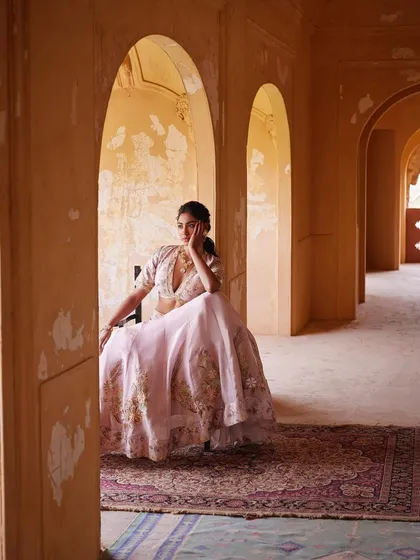 A campaign shot for Wedding Asia, capturing a quiet moment of reflection. The model is styled in a delicate pink floral lehenga, seated within a rustic arched hallway to create a soft, romantic, and picturesque scene.