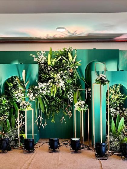 A modern stage design with a bold green backdrop, featuring geometric cutouts, gold stands, and an explosion of white and green floral arrangements.
