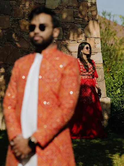 An artistic portrait using foreground blur to create depth. The focus is on the bride in her beautiful red lehenga, with the groom out of focus in the front.