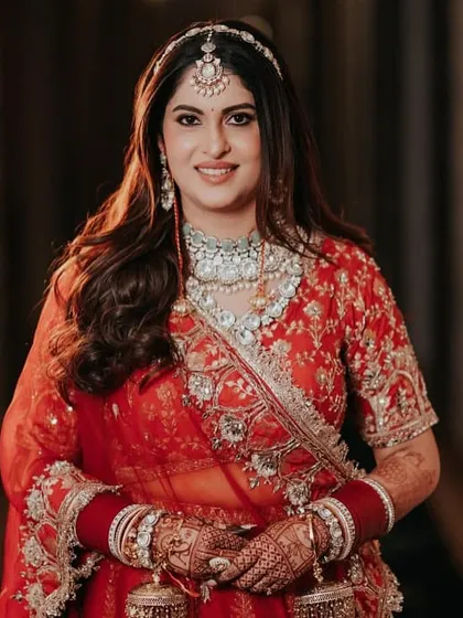 A classic, forward-facing bridal portrait. The soft lighting highlights her intricate jewelry and the rich color of her red lehenga, capturing her calm and radiant beauty just before the ceremony.