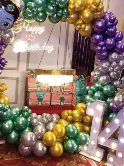 A unique and colorful Krishna-themed birthday decoration. The balloon arch uses purple, green, gold, and silver balloons, with cutouts of Little Krishna and a light-up number '14'.
