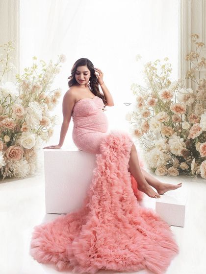A dreamy portrait of a mom-to-be seated amidst a beautiful floral arrangement. The soft pink ruffled gown cascades around her, creating a romantic and whimsical scene.