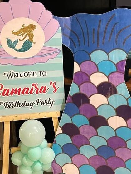 A personalized welcome sign for a mermaid-themed first birthday party. This custom easel stand features a mermaid in a clamshell and is paired with a large mermaid tail cutout, setting the theme right from the entrance.