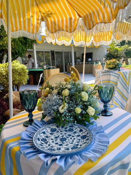 An outdoor table setting with a yellow and white striped umbrella, a blue and white striped tablecloth, and a beautiful floral centerpiece.