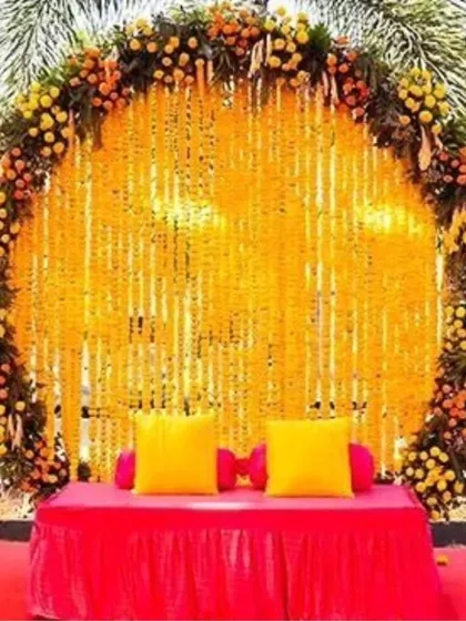 A vibrant outdoor Haldi photo booth with a circular backdrop of marigold strings. The bright pink seating provides a beautiful pop of color against the yellow flowers.