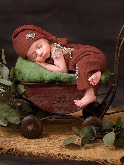 This baby is posed in a miniature vintage-style cart, surrounded by eucalyptus leaves on a burlap surface, showcasing our detailed rustic setups.