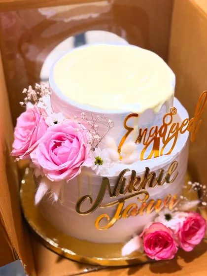 This two-tier engagement cake features a white chocolate drip, fresh pink roses, and custom gold acrylic name toppers for the happy couple.