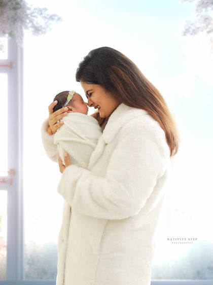 A mother shares a quiet moment with her newborn against a bright, wintery window backdrop. The soft, all-white theme gives the image a dreamy, serene quality.
