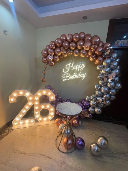 This is a duplicate of ID 37, 151, 158, 203, 247. A stylish half-arch balloon ring for a 26th birthday. This home decoration uses chrome rose gold and silver balloons, a neon sign, and marquee numbers.