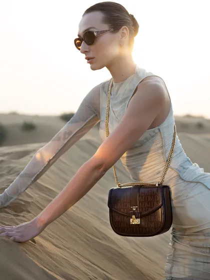 A sunset shot from the Da Milano desert campaign. The model is styled in a semi-sheer, ruched dress, with the warm light catching the texture of both the garment and the brown leather crossbody bag.