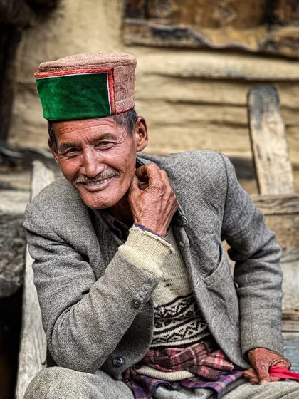 A smiling man in a traditional Himachali cap sits on a wooden porch. His warm expression is a testament to the friendly nature of the people in these mountains.