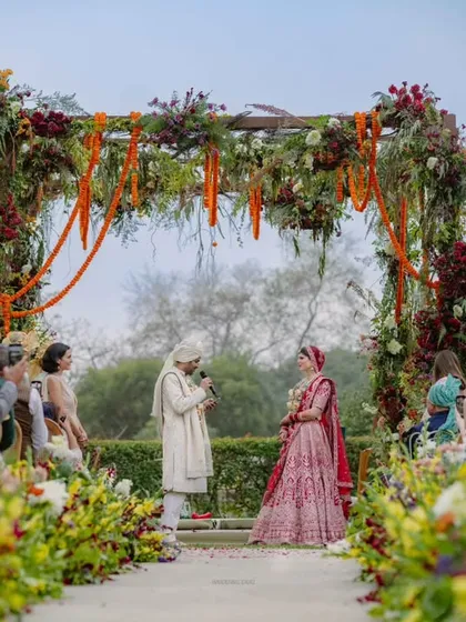 The couple exchanges vows under a lush floral mandap. The aisle is lined with wildflowers, and grand chandeliers hang overhead, crafting a perfect moment where love is sealed.