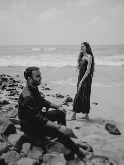 A moody black and white portrait on a rocky beach. The composition, with the couple in different planes, creates an artistic and contemplative feel.