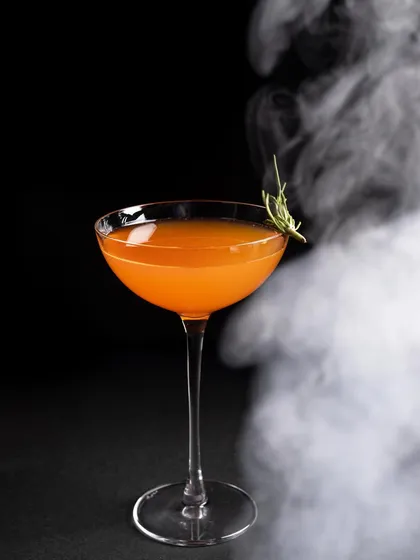 An orange cocktail with a sprig of rosemary, surrounded by a dramatic swirl of smoke. This shot is all about creating atmosphere and a sense of mystery.
