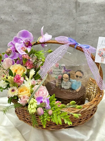 I create personalized gifts that tell a story, like this anniversary hamper. It features a Belgium chocolate cake with custom hand-made caricatures of the couple, paired with a beautiful fresh flower basket.