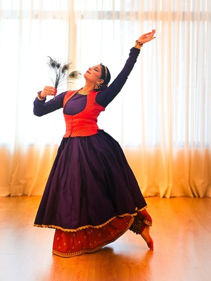 A dynamic pose from a Janmashtami-themed photoshoot, reaching upwards in a gesture of offering to Krishna. The peacock feather is a classic symbol of the deity.