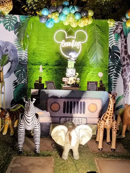 An immersive jungle safari theme for a child's birthday. This detailed cake-cutting area features a faux grass backdrop, a custom neon name sign, a jeep-shaped table, and life-sized inflatable safari animals like a giraffe, zebra, and elephant.