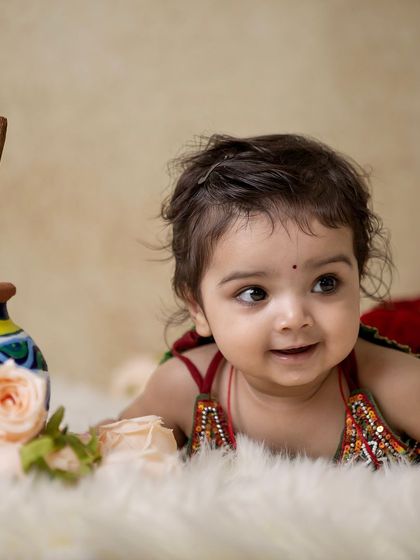 A candid shot of a baby girl in her Radha costume, lying on a soft fur rug. The focus is on her curious gaze and the traditional props beside her.