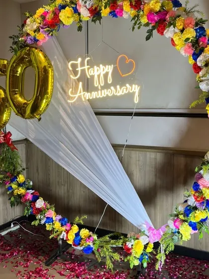 A grand 50th-anniversary decoration featuring a large ring covered in colorful artificial flowers, a 'Happy Anniversary' neon sign, and '50' foil balloons.